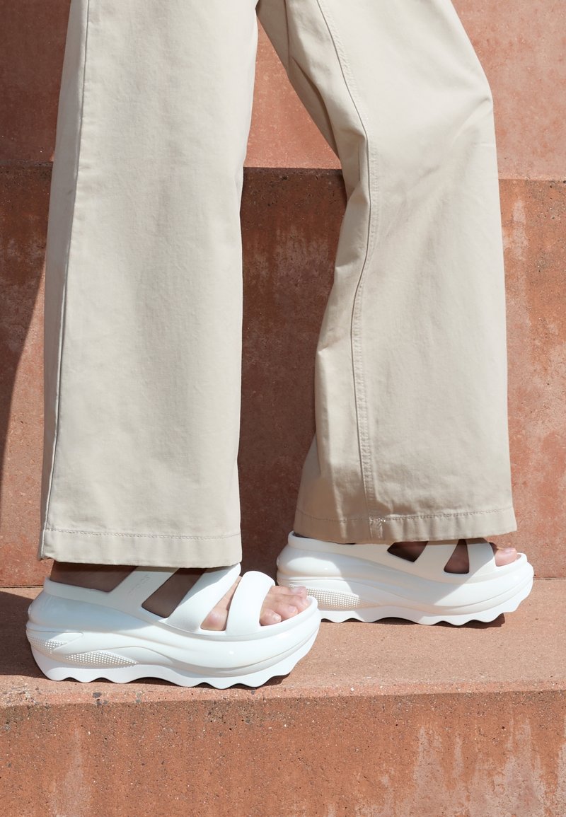 Pantalon beige à jambes larges associé à des sandales compensées blanches épaisses avec de larges brides, portées sur des marches de couleur terre cuite.