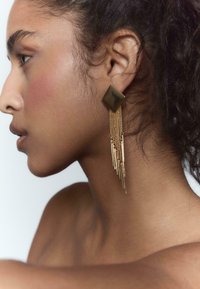 Orecchini in tono oro con un pezzo superiore a forma di diamante, dotati di catene pendenti e accenti metallici allungati, su uno sfondo neutro.