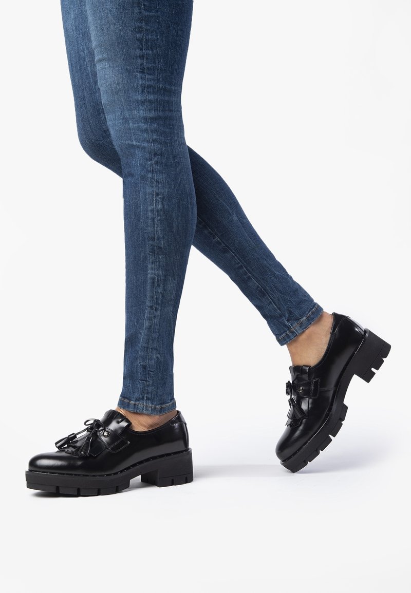 Mocassins en cuir verni noir avec une semelle plateforme épaisse, ornés d'un nœud à l'avant et d'une texture lisse et brillante.