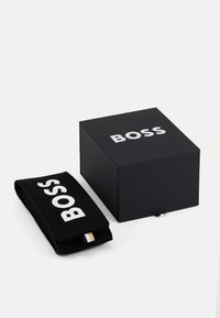 Juodas dėžutė su balta "BOSS" logotipu, kuriame yra juodas sukučiukas su logotipu ir maža audinio etiketė. Kompaktiška ir stačiakampė.