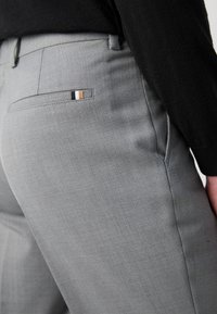 Grå skreddersydde bukser med strukturert stoff, slim fit, og en liten baklomme med stripete detalj i svart, hvitt og beige.