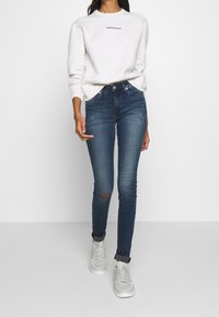 Mujer vistiendo un sudadera blanca de Calvin Klein, jeans ajustados azules rasgados y zapatillas blancas, de pie contra un fondo blanco liso.