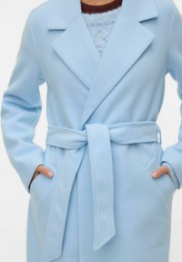 Hele sinine ümberseotav mantel pehmest kangast, vöösil, suurte kraede ja küljetaskutega. Selle all tekstuurse kudumiga top.