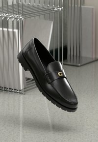 Mocassin en cuir noir avec une semelle épaisse et un détail de boucle dorée, flottant au-dessus d'un sol gris tacheté près de supports métalliques tenant des objets plats.