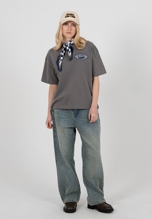 Woman wearing a beige cap, checkered scarf, gray oversized t-shirt with a logo, loose blue jeans, and dark brown shoes, standing against a plain background.