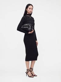 Femme en robe midi noire à manches longues tenant un sac à main en cuir noir, portant des sandales à talons hauts noires à brides, sur fond blanc.