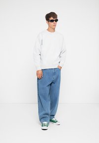 Hanorac gri, blugi largi albaștri și adidași cu spirală verde. Modelul poartă ochelari de soare negri, stând în fața unui fundal alb.