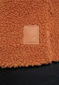 Gros plan sur un tissu polaire orange texturé avec une petite pièce en cuir cousue comportant un logo minimaliste embossé.