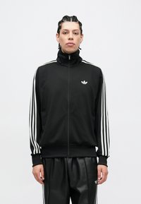 Čierna bunda na zips s vysokým golierom, biele tri pruhy na rukávoch a malým bielym logom na hrudi, v kombinácii s čiernymi nohavicami.