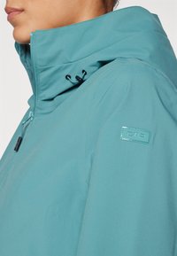Teal værbestandig jakke med høy krage, justerbar snøring og en logopatch. Glatt tekstur og glidelåsdetalj.