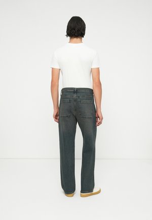 Homme aux cheveux noirs portant un t-shirt blanc, un jean gris foncé et des chaussures marron clair, debout face à un mur blanc.