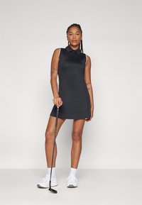 Svart ärmlös golfklänning med polo-krage, sidofickor och kontrasterande vita detaljer. Modell som håller i en golfklubba. Vita skor på fötterna.