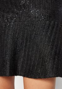 Jupe noire en tricot avec des fils métalliques brillants subtils tissés dans le tissu, portée sur des jambes nues contre un fond neutre.