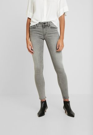 Person trägt ausgebleichte graue Skinny-Jeans, schwarze Stiefeletten mit kurzen Absätzen und ein lockeres weißes Hemd mit Knöpfen.