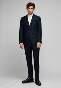 Marineblazer mit strukturiertem Stoff und Reverskragen, kombiniert mit einem cremefarbenen Rollkragenpullover, marineblauen Hosen und schwarzen Schnürschuhen.