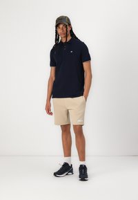Navy polo shirt with white accents, beige shorts with logo, and black athletic shoes. Model wears a camouflage cap and has braided hair.