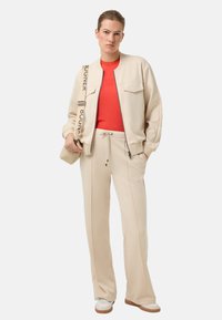 Crèmewitte bomberjack met twee opvallende zakken, gecombineerd met een rood t-shirt en beige broek met trekkoord. Model draagt witte sneakers.