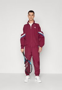 Burgundijas krāsas treniņtērps ar baltām un ziliem svītrām, jaku ar rāvējslēdzēju un elastīgām aprocēm. Modelis tur rokās melnu tenisa raketi un valkā baltas kedas.