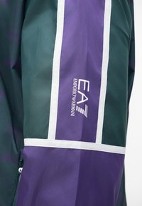 Zielono-fioletowy wiatrówka z białymi akcentami, z logo "EA7 Emporio Armani" na rękawie i teksturowanym materiałem. Zamek błyskawiczny.