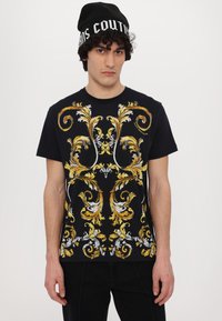 T-shirt preta com um intrincado padrão barroco em amarelo e branco, gola redonda, mangas curtas. Modelo usa um gorro preto com texto branco.