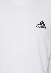 Valkoinen puuvillaiset T-paita, jossa on pyöreä kauluskanna ja pieni musta Adidas-logo vasemmassa rinnassa. Sileä rakenne, suora leikkaus.