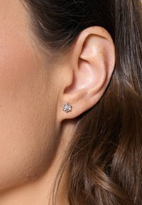 Okrągłe kolczyki-sztyfty z diamentami osadzone w metalowej obrączce w srebrnym odcieniu, z kwiatowym wzorem i małymi kamieniami akcentującymi wokół centralnego diamentu.