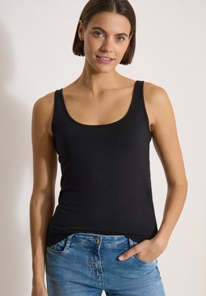 Femme aux cheveux courts bruns portant un débardeur noir et un jean bleu, une main dans la poche, souriante à la caméra.