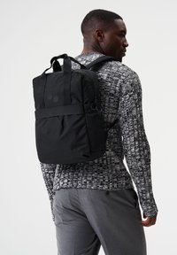 Schwarzer Rucksack mit gepolsterten Trägern, rechteckiger Form und minimalistischen Design. Verfügt über eine frontale Reißverschlusstasche und dezente Logo-Akzente.