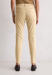 Persoon in beige slim-fit broeken en donkerbruine leren instappers, staand tegen een effen witte muur.