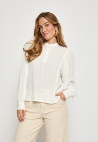 Balta, ilga rankovėmis mygtukinė palaidinė su apykakle, su tekstūruotu langelių raštu ir laisvu kirpimu, derinama su šviesiai bežė kelnėmis.