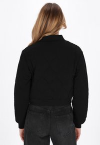 Frau mit langen Haaren, die eine schwarze gesteppte Jacke und schwarze Jeans trägt, von hinten vor einem weißen Hintergrund gezeigt.