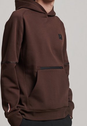 Persona que lleva una sudadera con capucha marrón con un bolsillo delantero con cremallera y una parche negro en el pecho, manos en los bolsillos laterales.
