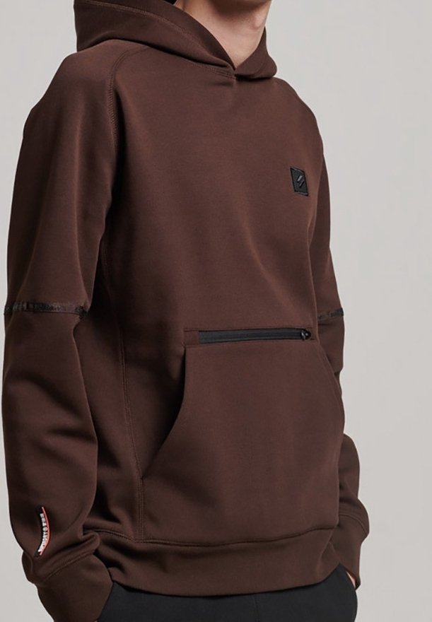 Personne portant un sweat à capuche marron avec une poche avant zippée et un patch noir sur la poitrine, mains dans les poches latérales.
