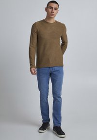 Maglione a maniche lunghe marrone in maglia con scollo rotondo, abbinato a jeans di denim azzurri e sneakers nere. Texture liscia, design aderente.