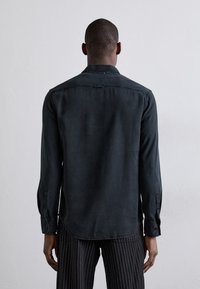 Camicia scura a maniche lunghe in tessuto morbido con colletto a bottoni, caratterizzata da un'orlo curvo e polsini con bottone, vista da dietro.