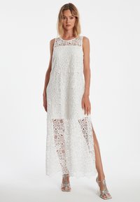 Weißes Maxikleid aus Spitze mit floralem Muster, Cut-out-Design und seitlichen Schlitzen. Hat einen runden Ausschnitt und ein ärmelloses Design.