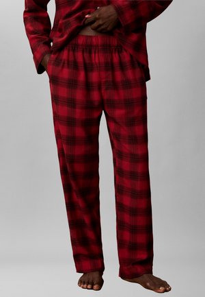 Mand iført røde og sorte ternede pyjamasbukser med elastisk talje og matchende top, stående barfodet mod en ensfarvet baggrund.