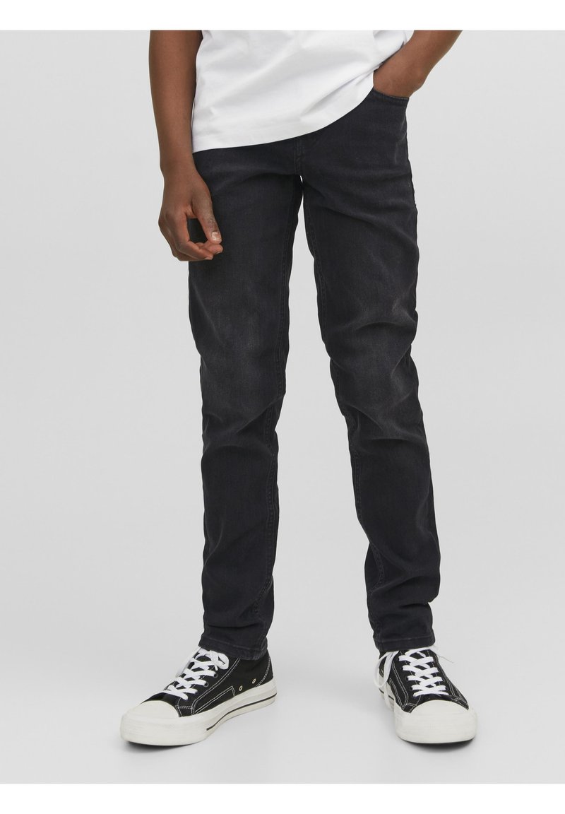 Homme portant un jean noir, un t-shirt blanc et des baskets noir et blanc, debout avec une main dans la poche devant un fond uni.