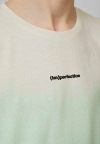 Koszulka z krótkim rękawem w kolorze kremowym z gradientowym efektem zieleni. Posiada haftowany napis "(im)perfection" w kolorze czarnym. Wykonana z bawełny, okrągły dekolt.