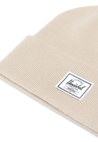 Beige strikket beanie med ribbet tekstur og foldet opretræk. Har et rektangulært sort og hvidt mærkelabel, som er præsenteret tydeligt.