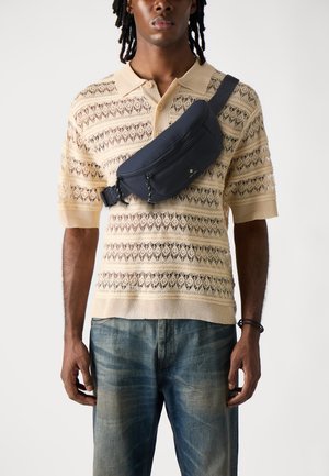 Navy crossbody bag with zippered pocket and adjustable strap worn over a beige patterned knitted polo shirt with a loose fit and short sleeves.