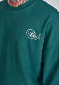 Sudadera de cuello redondo en tono verde azulado, hecha de una mezcla de algodón, con un logo blanco en el lado izquierdo del pecho. Textura suave con cuello y puños acanalados.