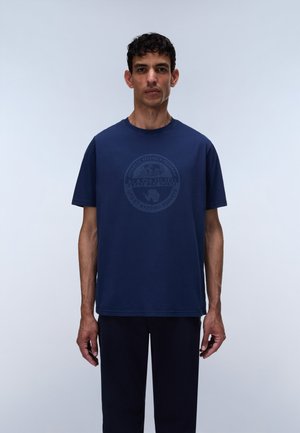 Homme debout sur un fond uni, portant un t-shirt bleu marine avec un faible emblème circulaire et un pantalon sombre.