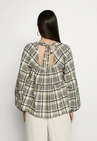 Blouse à manches longues avec un trou de serrure dans le dos, présentant un motif carré vert et noir, un design plissé et une fermeture nouée au niveau du col.