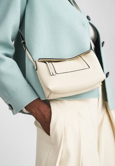 Bílá kožená crossbody taška se strukturovaným designem, tenkými černými obrysy a jedním nastavitelným popruhem, nošena pod světle modrým kabátem.