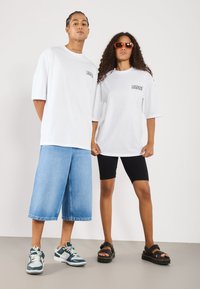 T-shirts blancs oversize avec "LEGENDS" imprimé sur le devant, associés à des shorts en denim à jambes larges et à des shorts de vélo noirs. Baskets décontractées et sandales.