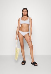 Conjunto de biquíni lilás claro com decote quadrado e alças finas, acompanhado de uma bolsa de mão amarela e branca às riscas e chinelos pretos.