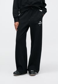 Μαύρο, φαρδύ παντελόνι φόρμας με ελαστική μέση και κορδόνι. Διαθέτει λευκό λογότυπο «Nike Athletics» στον αριστερό μηρό. Απαλό βαμβακερό υλικό.