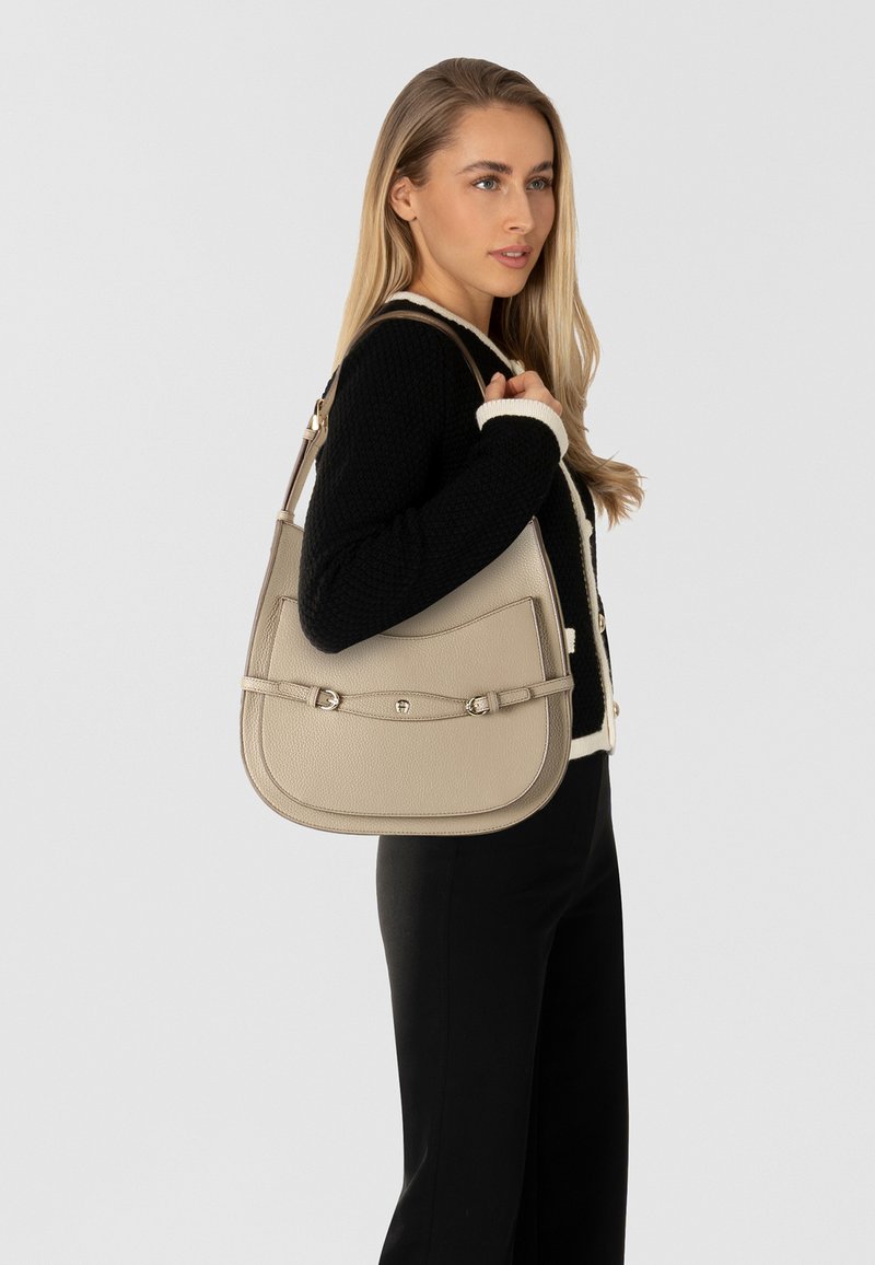 Beige leren schoudertas met een gladde textuur, gebogen vorm en decoratieve metalen accenten. Voorzien van een verstelbare riem.