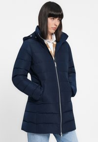 Giacca puffa blu marina con zip frontale, cappuccio e tasche laterali. Presenta un motivo a quadretti orizzontali e una texture morbida e liscia.